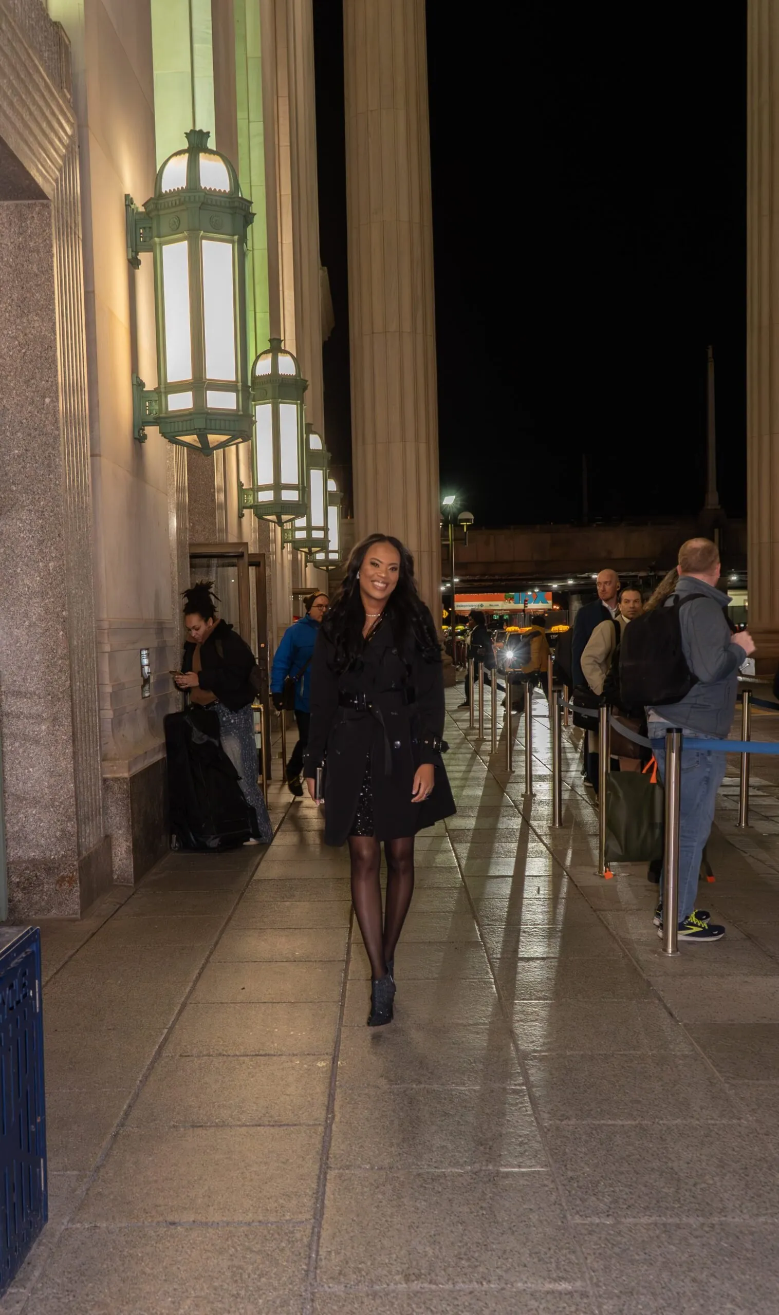 Rosa Allison walking towards the camera at night along a lantern-lit colonnade outside a grand stone building, smiling, in a black belted trench coat.
