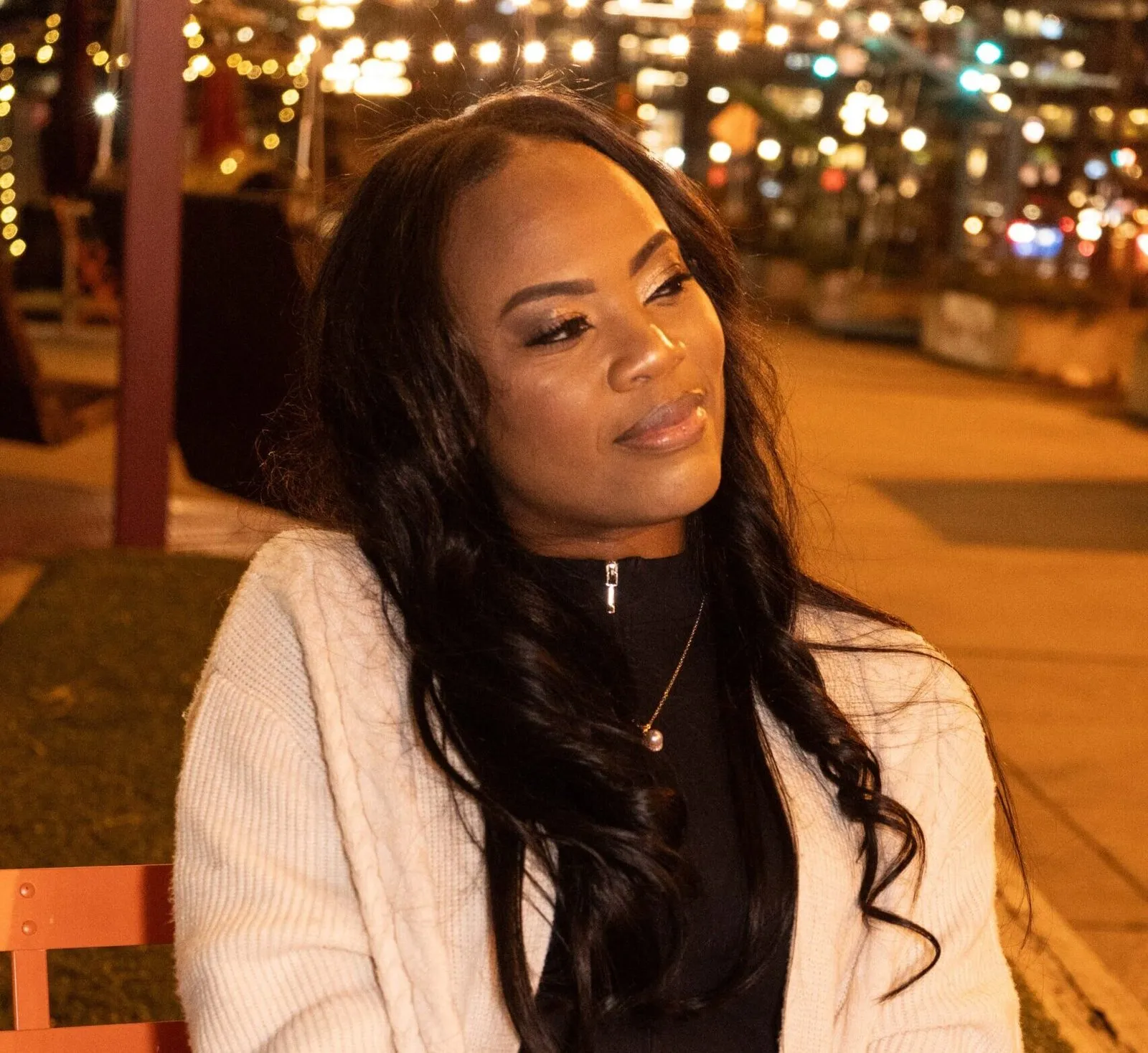 Rosa Allison seated outdoors in the evening in a cream cable-knit cardigan over a black top, looking away from the camera, with string lights and city lights blurred behind her.